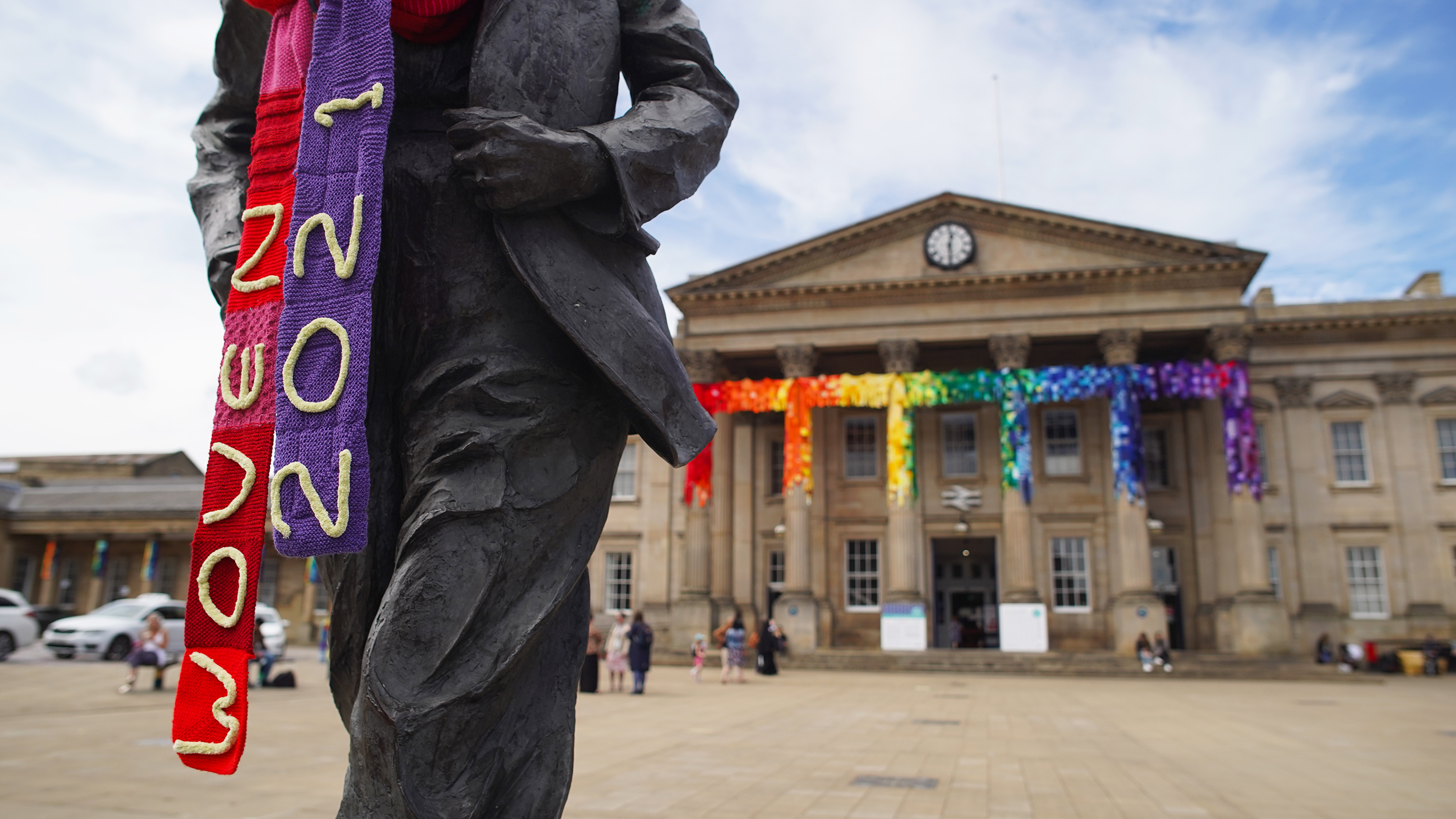 Front of Huddersfield Train Station in the background showing the knitted rainbow decoration on the pillars with a statue in the foreground wearing a knitted WOVEN2021 scarf