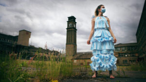 A woman in a dress made from face masks and bare feet in front of a building with a tower