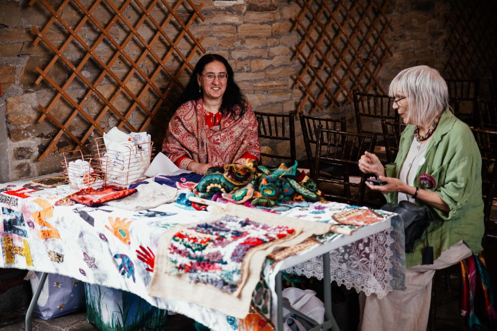 Two people sitting at a table displaying embroidery, knitted mandalas, textiltes and more