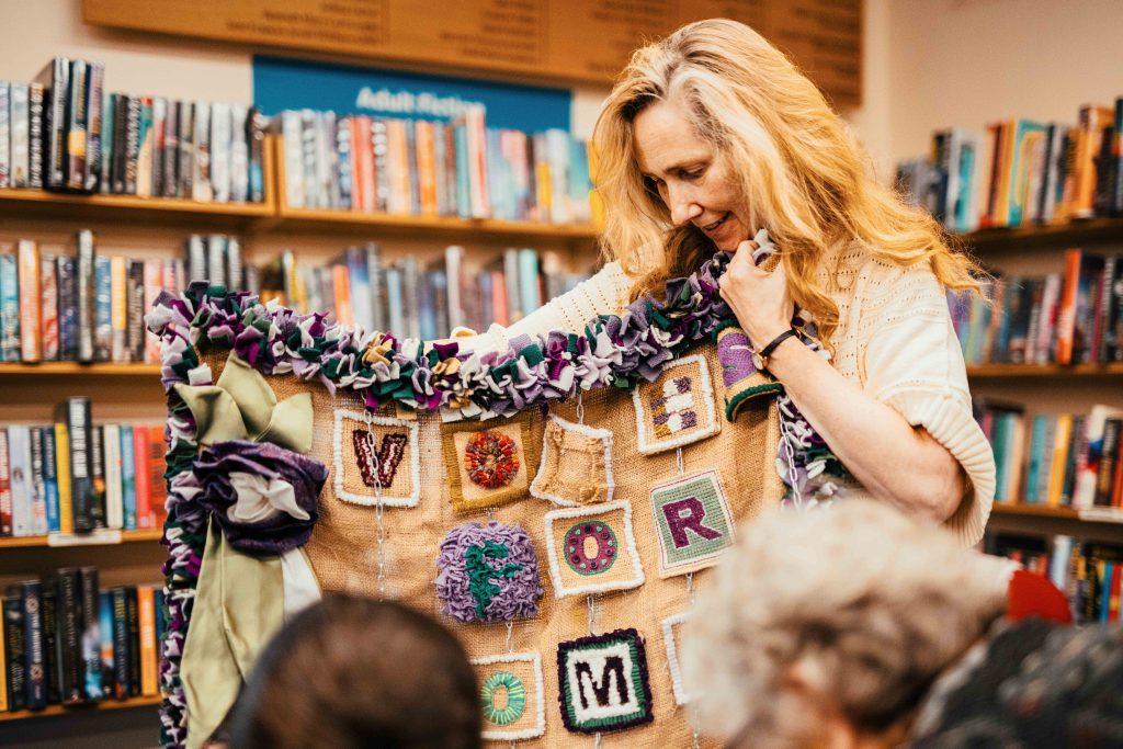 A person in a library setting holding up a textile banner with stitched squares saying votes for...