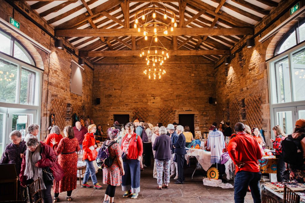 A large barn with chandeliers full of people and tables displaying textiles and garments