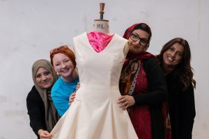 4 people smiling and peering from behind a calico dress on a dressmaker's mannequin