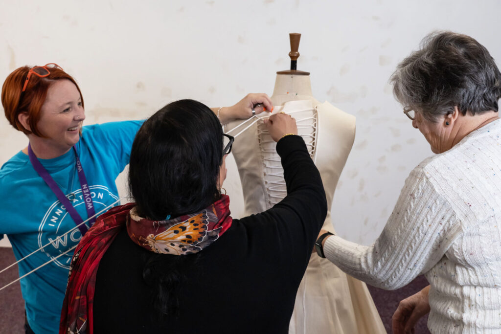 Three people pinning fabric onto a dress made of calico on a mannequin