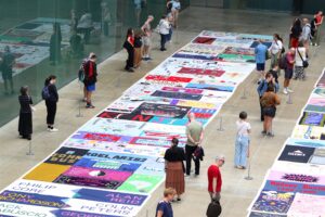 A large display of quilts in a gallery setting with people viewing them