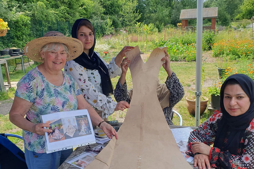A group of women holding up a large paper dress patterns in a garden
